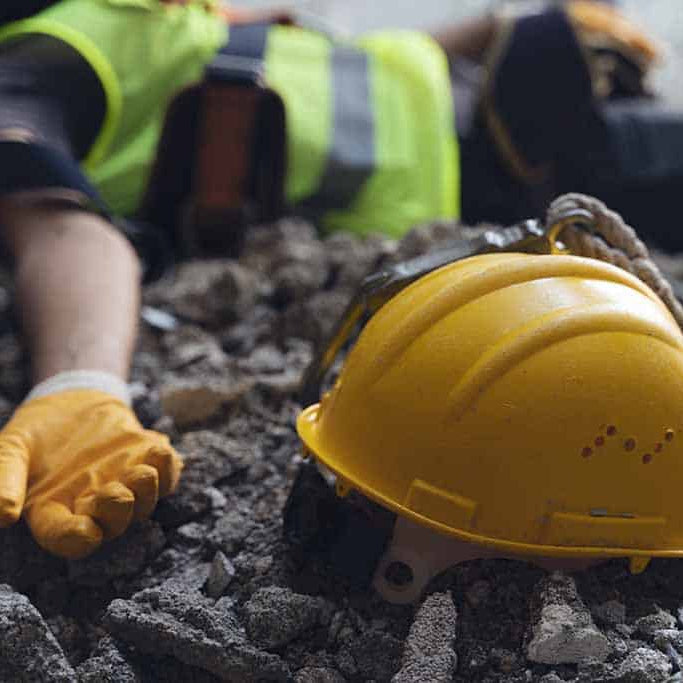 Yellow hard hat on rubble with a person wearing gloves and a high-visibility vest in the background.